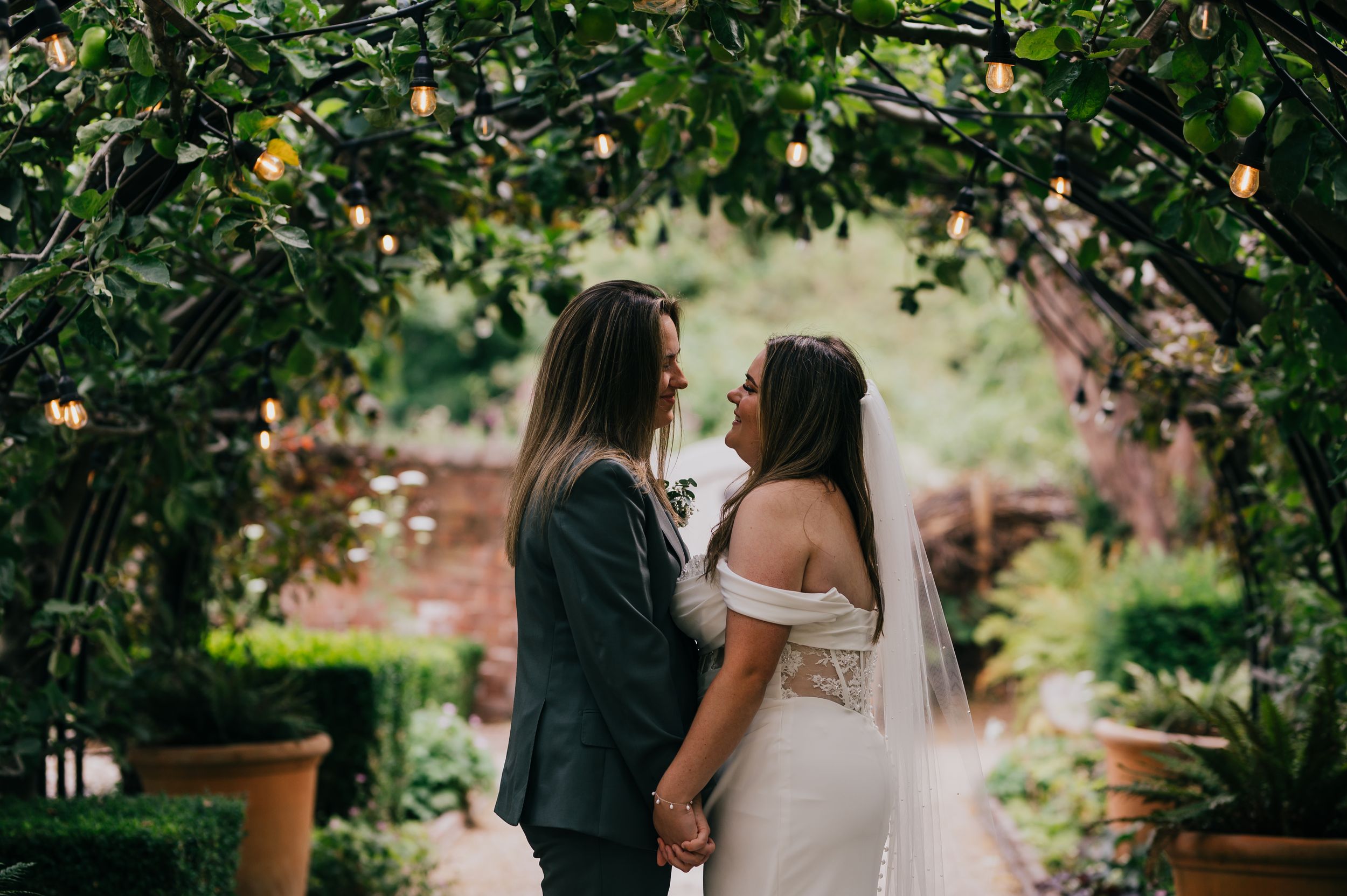 Two brides hold hands under a lush garden archway with string lights, sharing a romantic moment on their wedding day.
