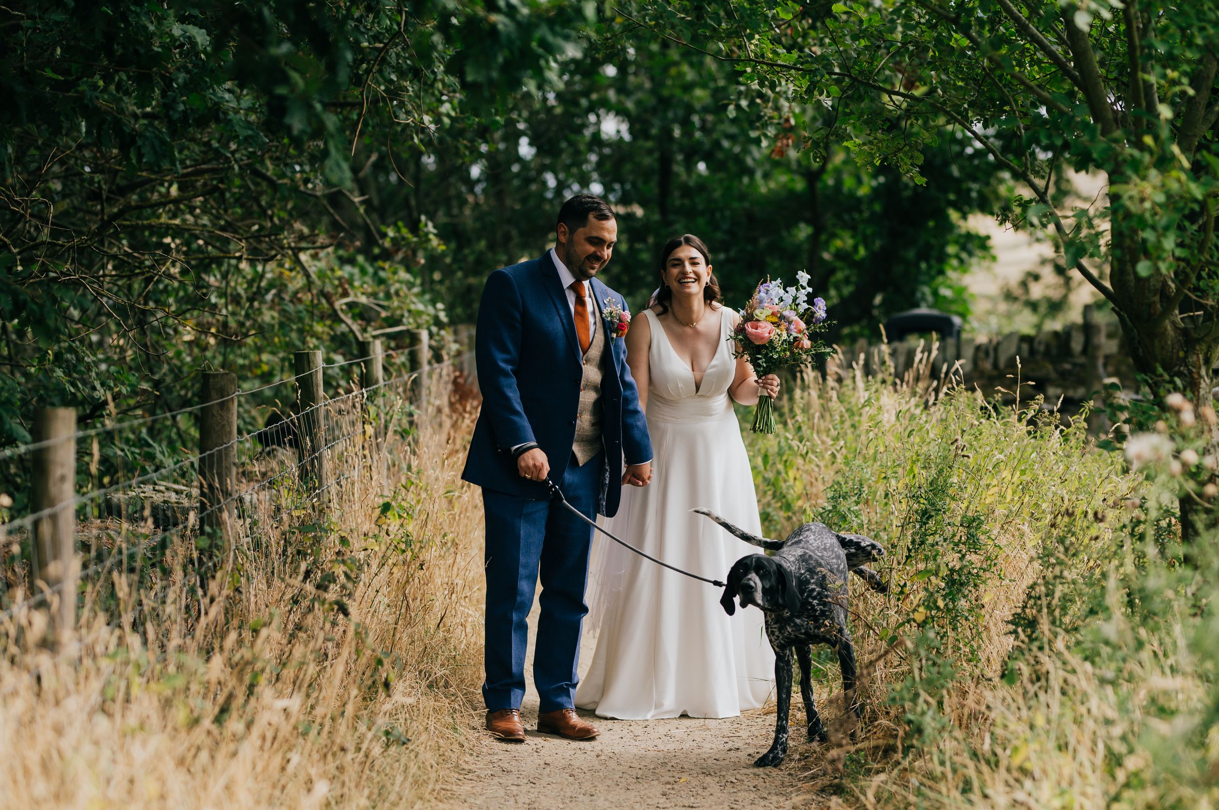 Bride and groom walking a black dog along a garden path on their wedding day.