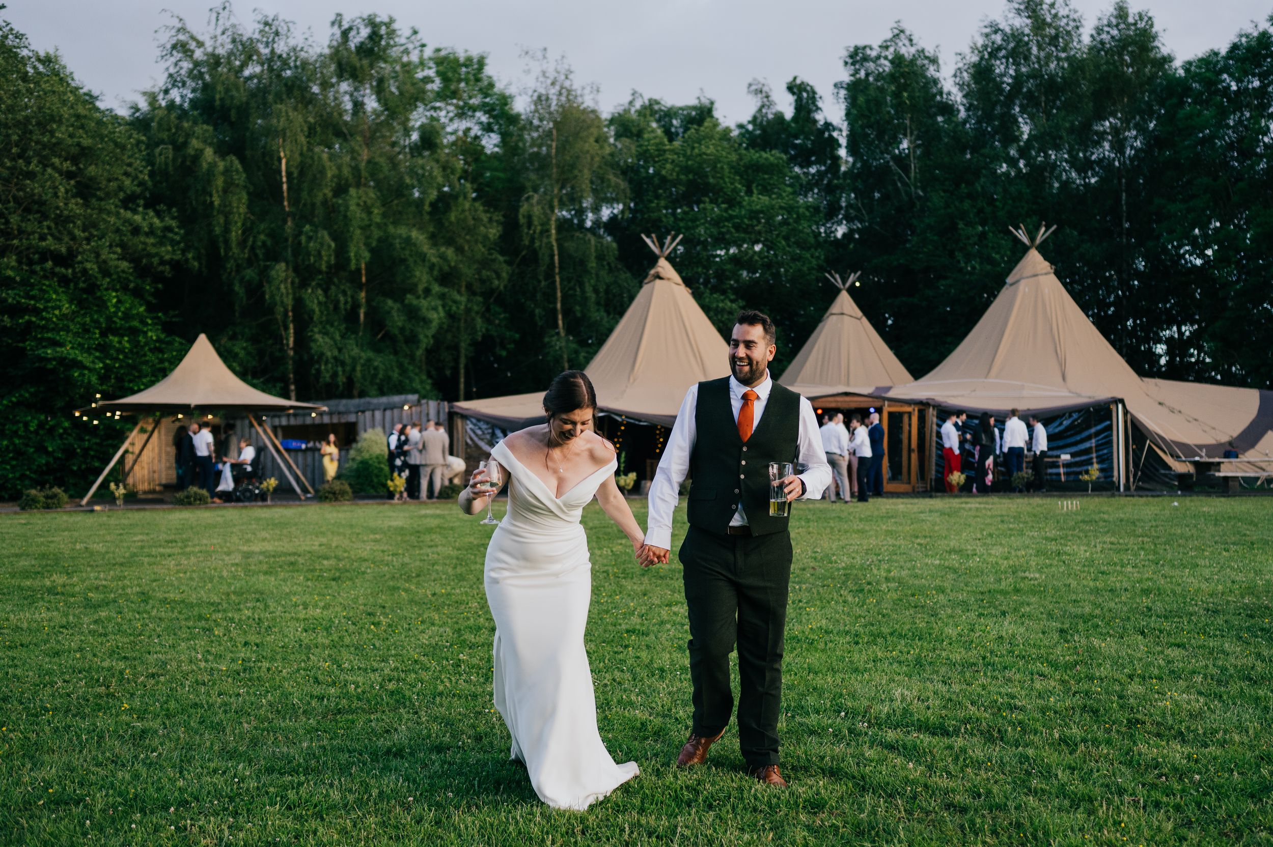 Bride and groom holding hands walking on green lawn with rustic tipi wedding venue tents in background.