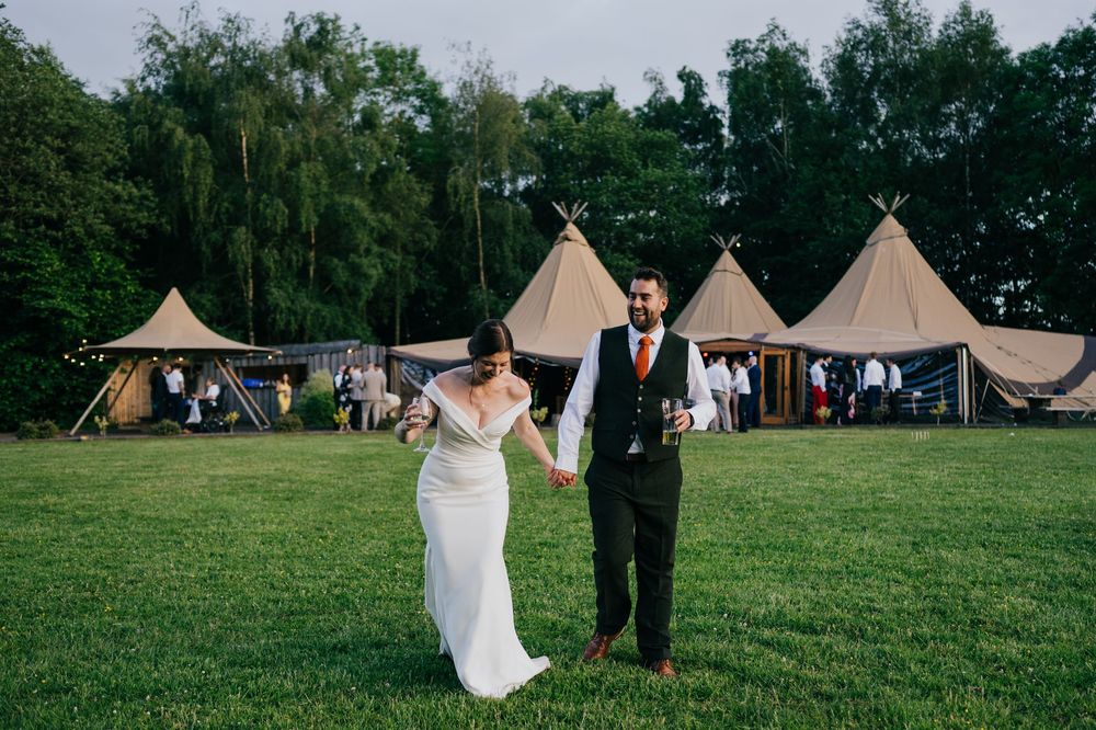 Bride and groom holding hands walking on green lawn with rustic tipi wedding venue tents in background.