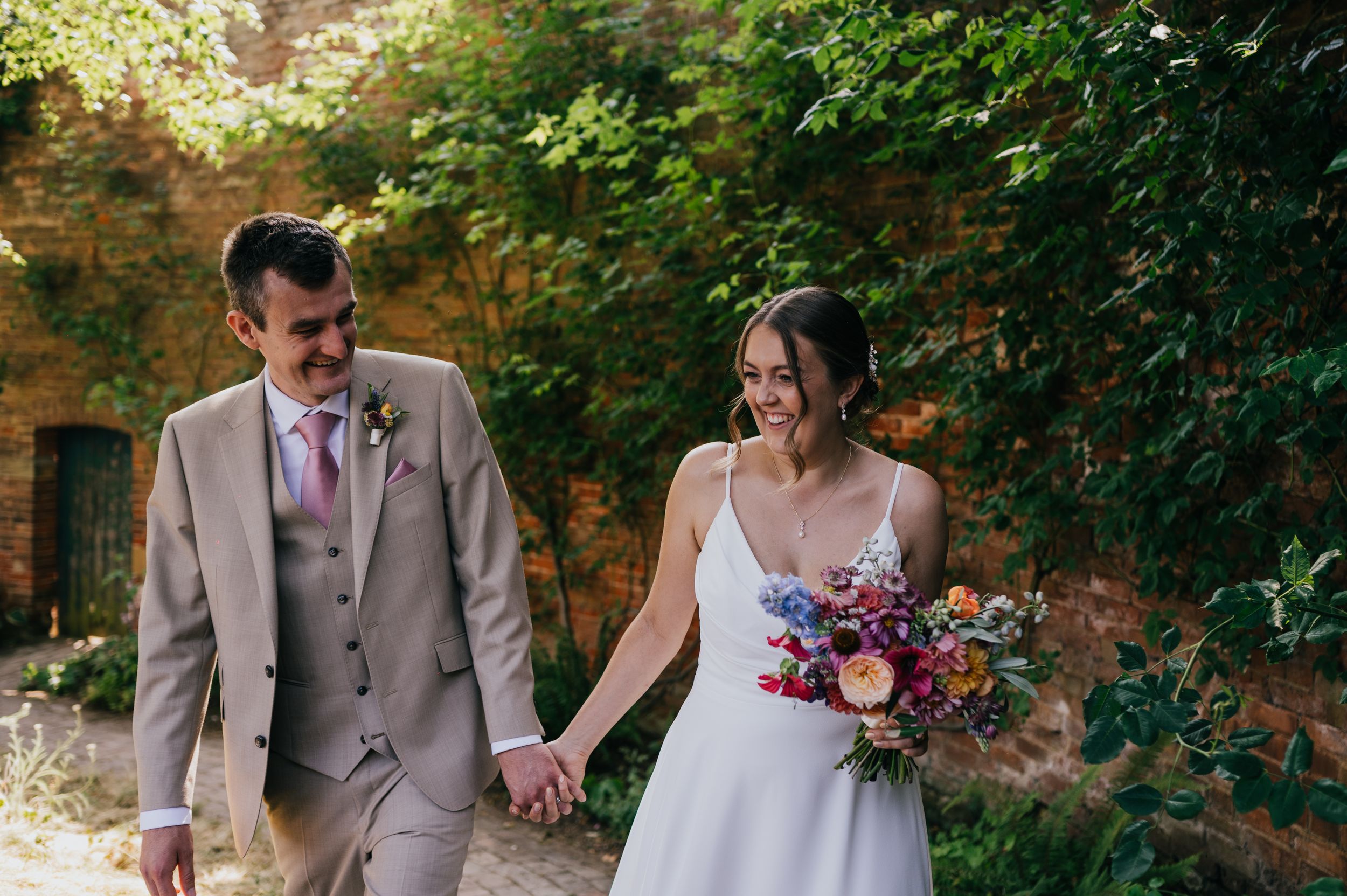 Bride and groom walk hand in hand outdoors, she holds a colorful bouquet, he wears a tan suit, surrounded by lush greenery.
