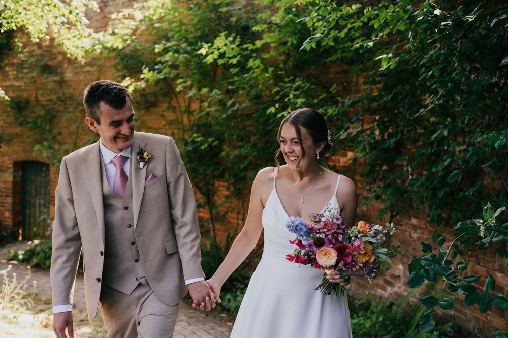 Bride and groom walk hand in hand outdoors, she holds a colorful bouquet, he wears a tan suit, surrounded by lush greenery.