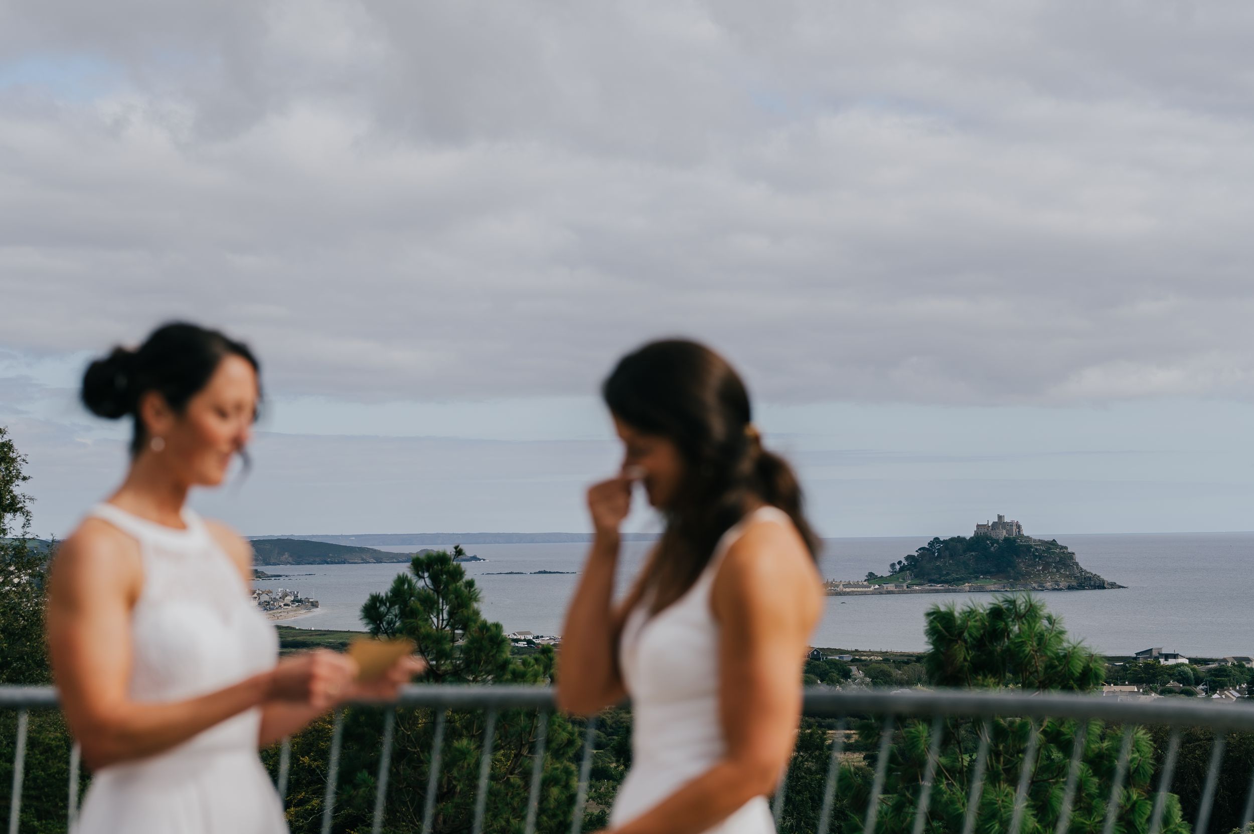 Two brides in white dresses stand on a balcony with St Michael's Mount visible in the background.