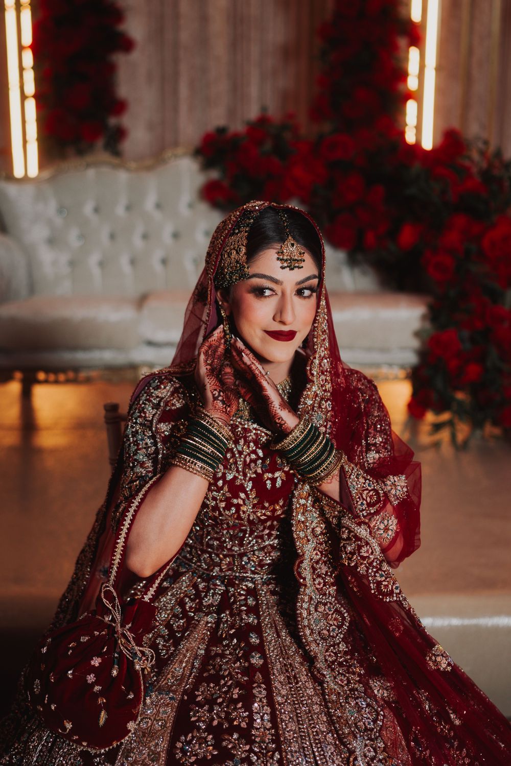 Bride in an intricately embroidered deep red lehenga poses against a backdrop of roses.