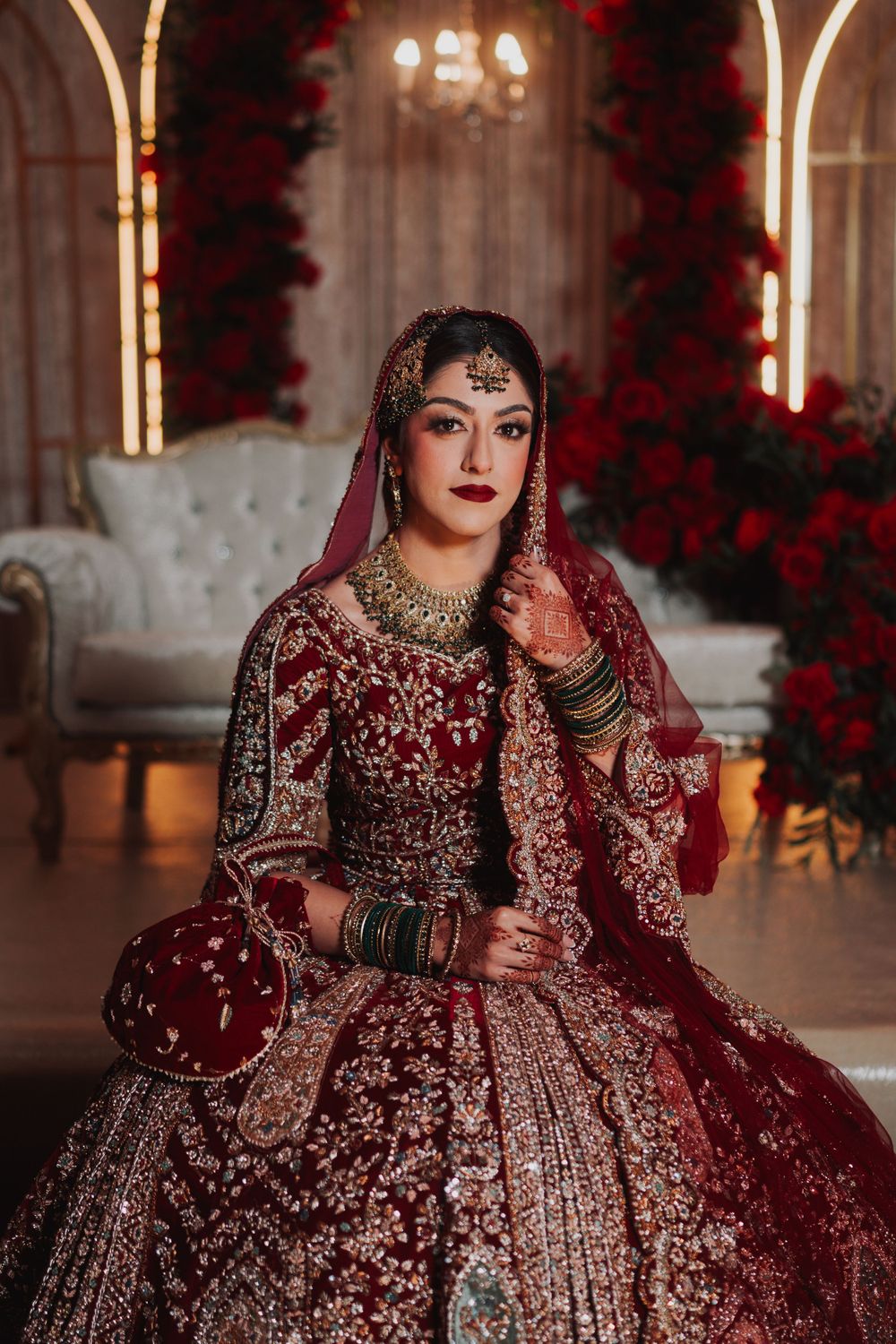 Bride in a heavily embellished burgundy lehenga sits on an elegant white sofa.