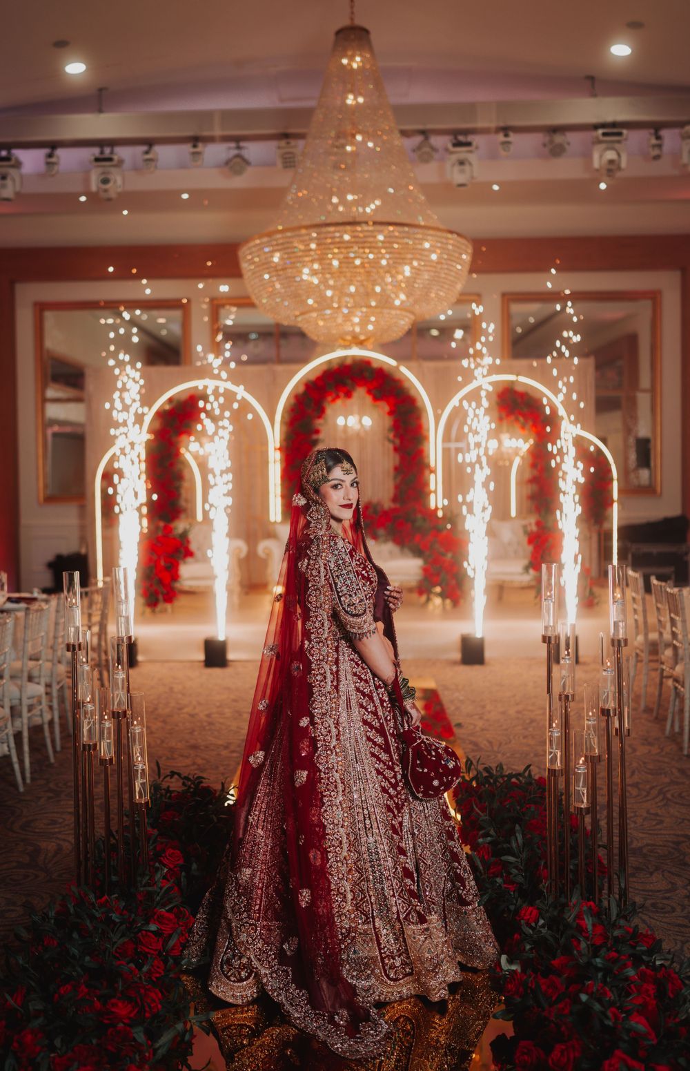 Bride in red embroidered lehenga poses under crystal chandelier with illuminated wedding arch backdrop.