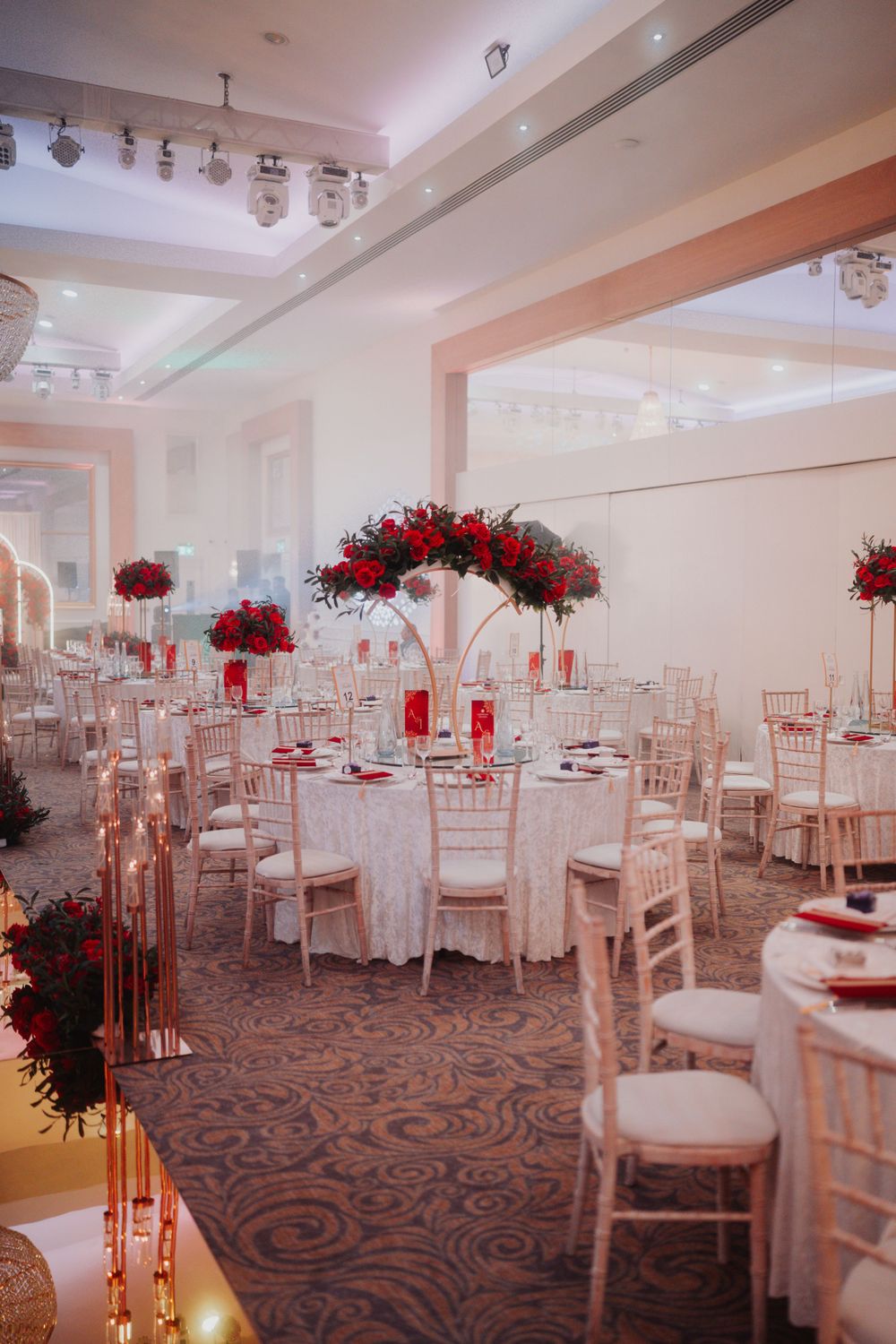 Elegant reception hall decorated with red rose centerpieces and white chiavari chairs.