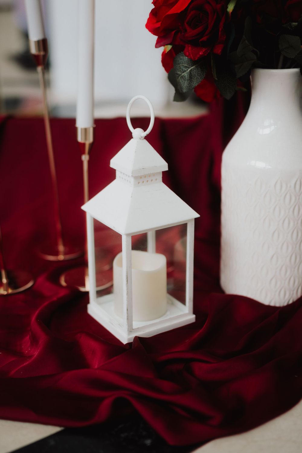 White decorative lantern with candle on red velvet tablecloth next to roses.