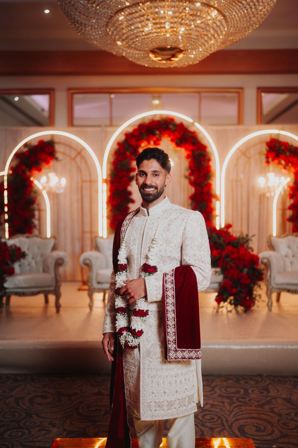Traditional South Asian groom in white sherwani and burgundy dupatta standing in front of decorated arches.