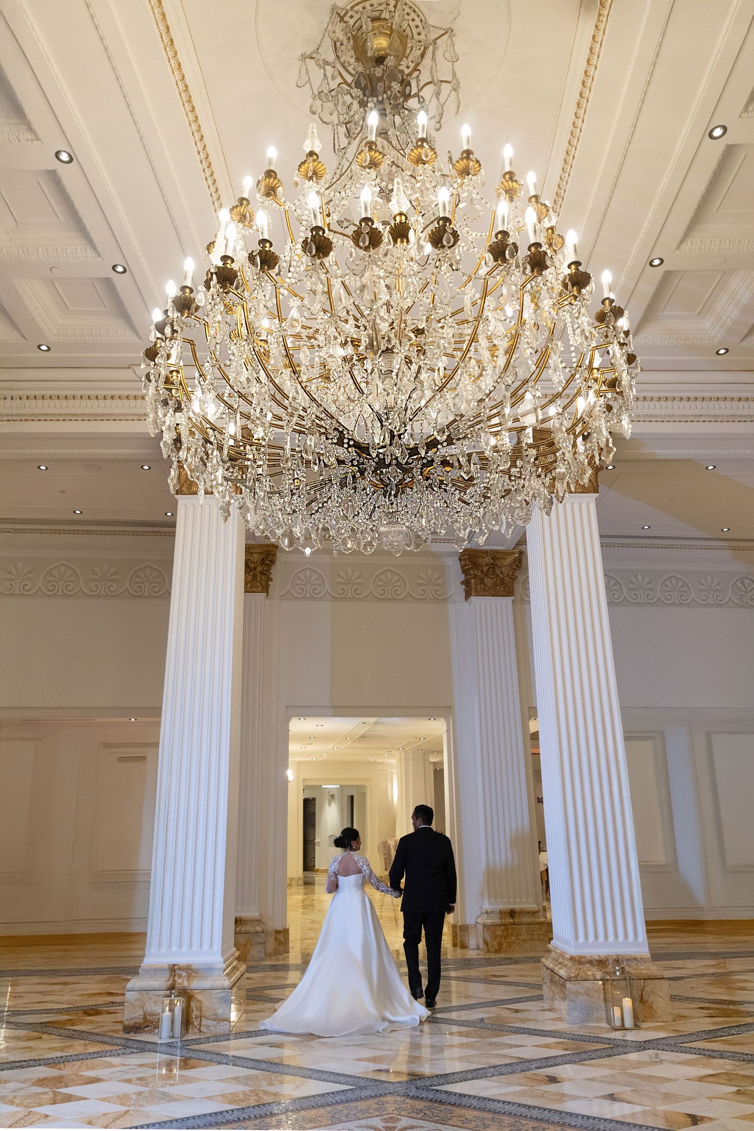 Bride and groom walking hand in hand beneath grand crystal chandelier in elegant ballroom.
