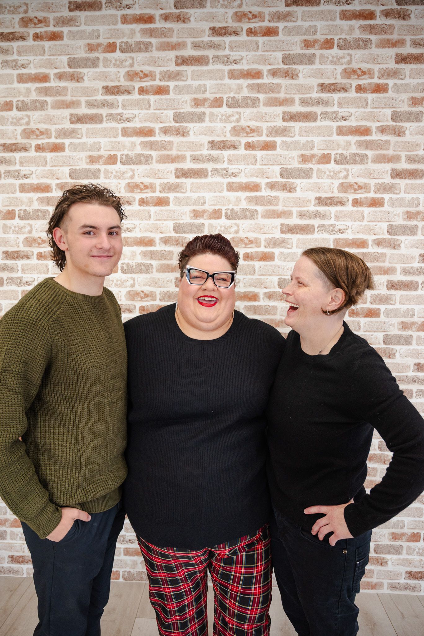 Three people smiling together against brick wall, wearing casual winter clothing indoors.