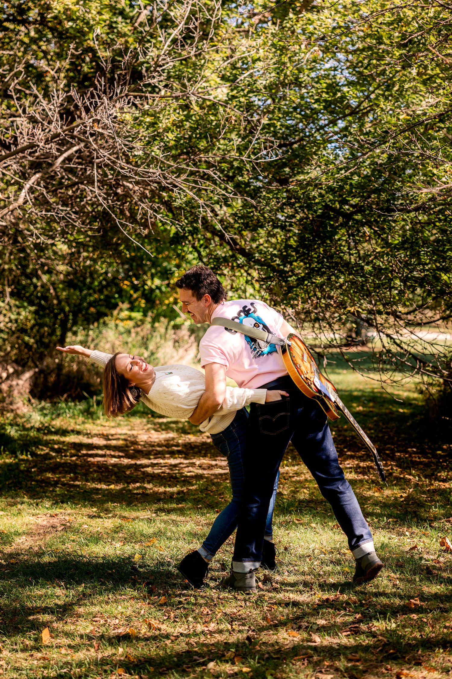 A couple dances together in a romantic dip pose under sunlit trees in an outdoor park setting in Novi Michigan