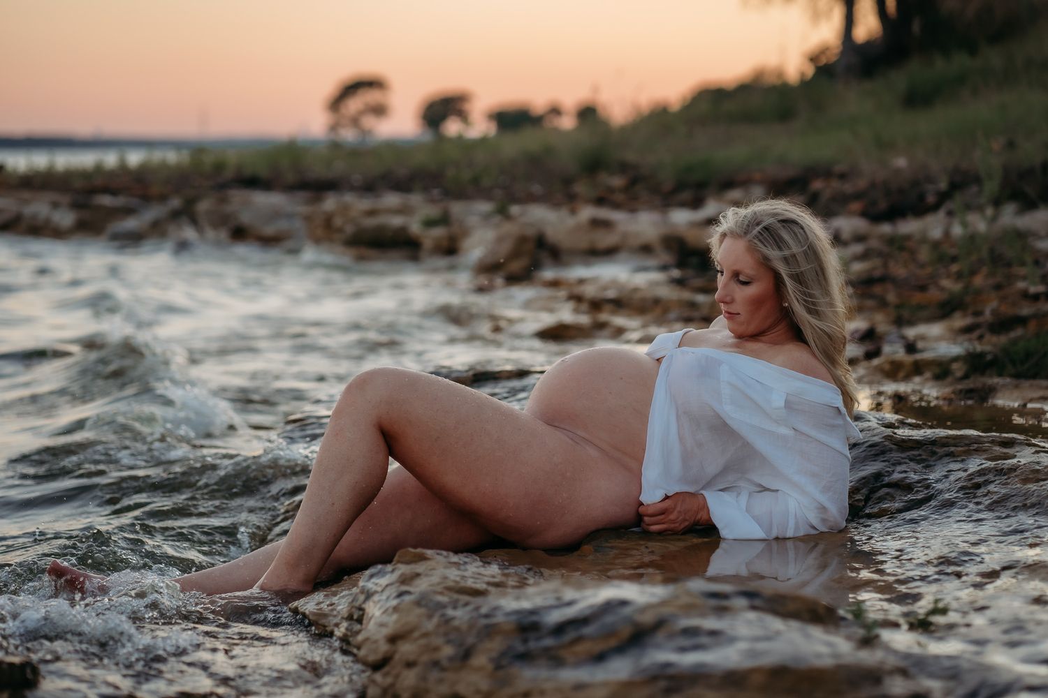 Maternity photo of an expectant mother in white dress reclining on rocky beach at sunset.