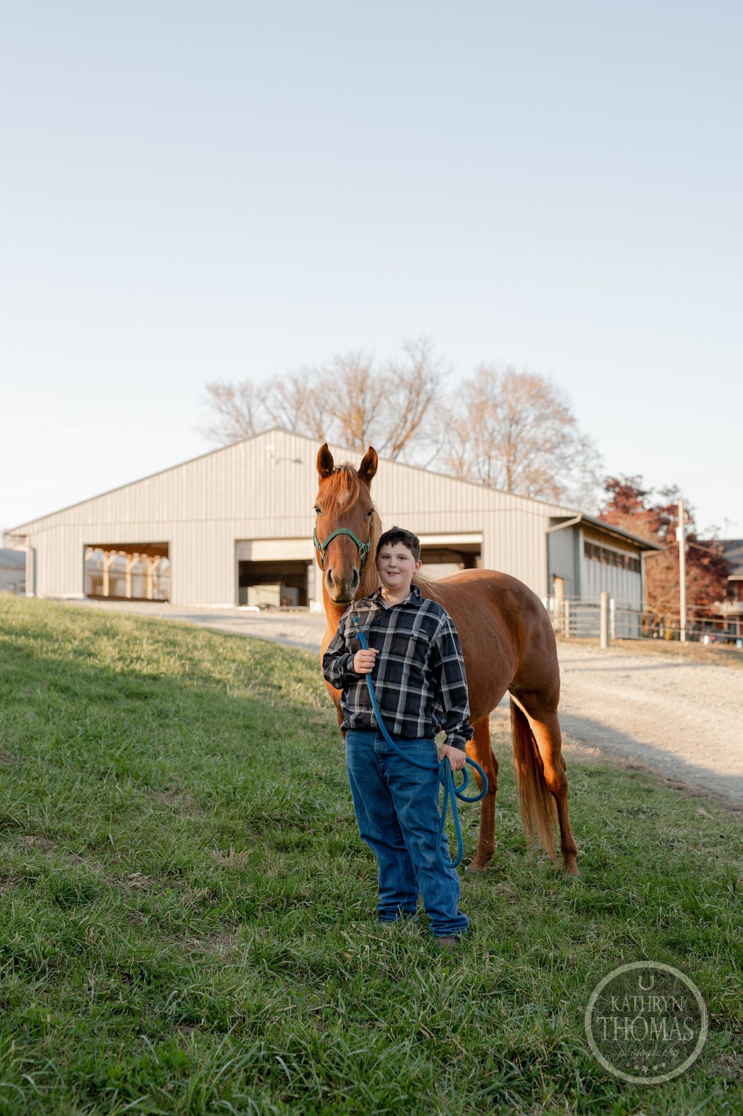 A young person stands with a chestnut horse outside a barn on a rural property.