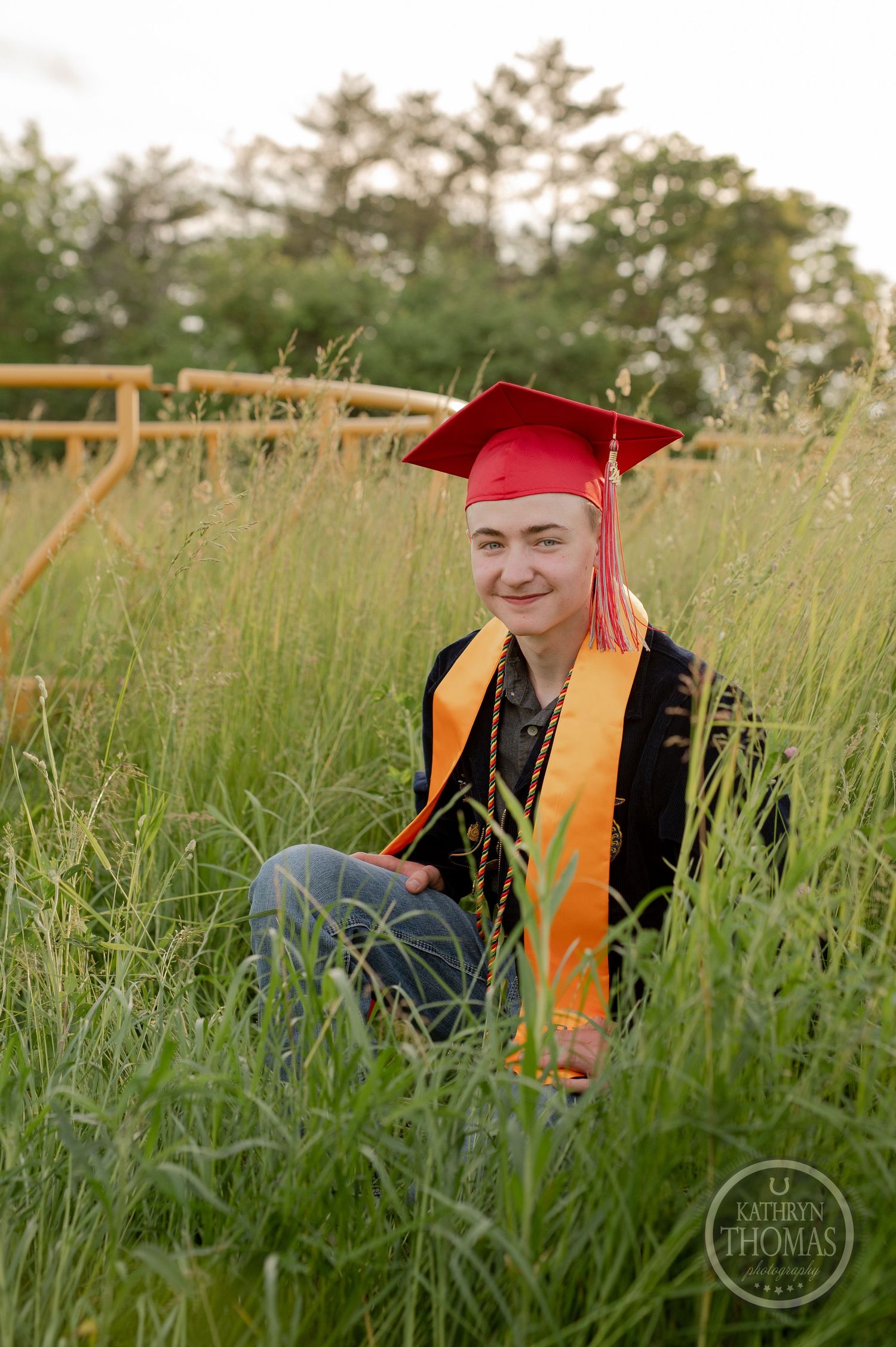 Graduate sits in tall grass wearing red cap, gold stole and black graduation gown.