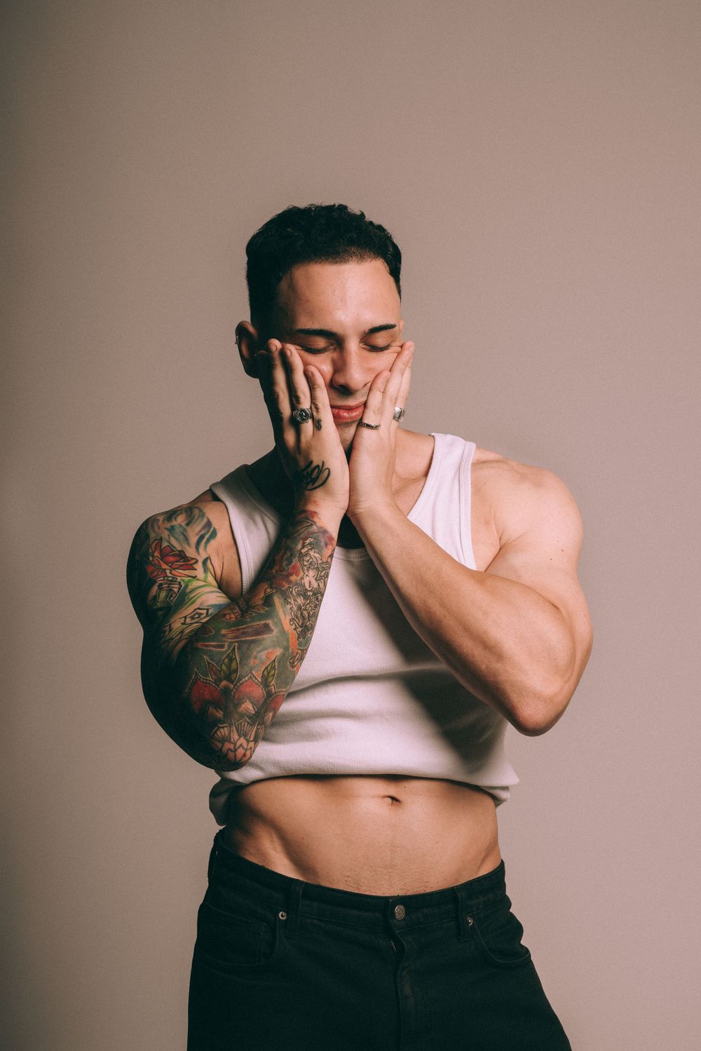 Athletic model in white tank top showing tattoo sleeve poses with hands on face.