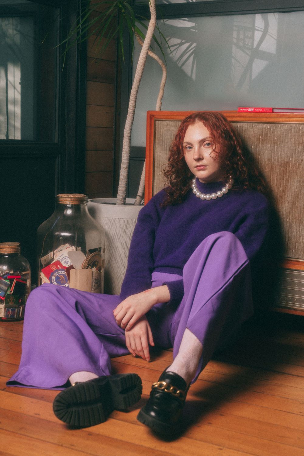 Model in purple sweater and pants sits casually in interior setting with houseplant.