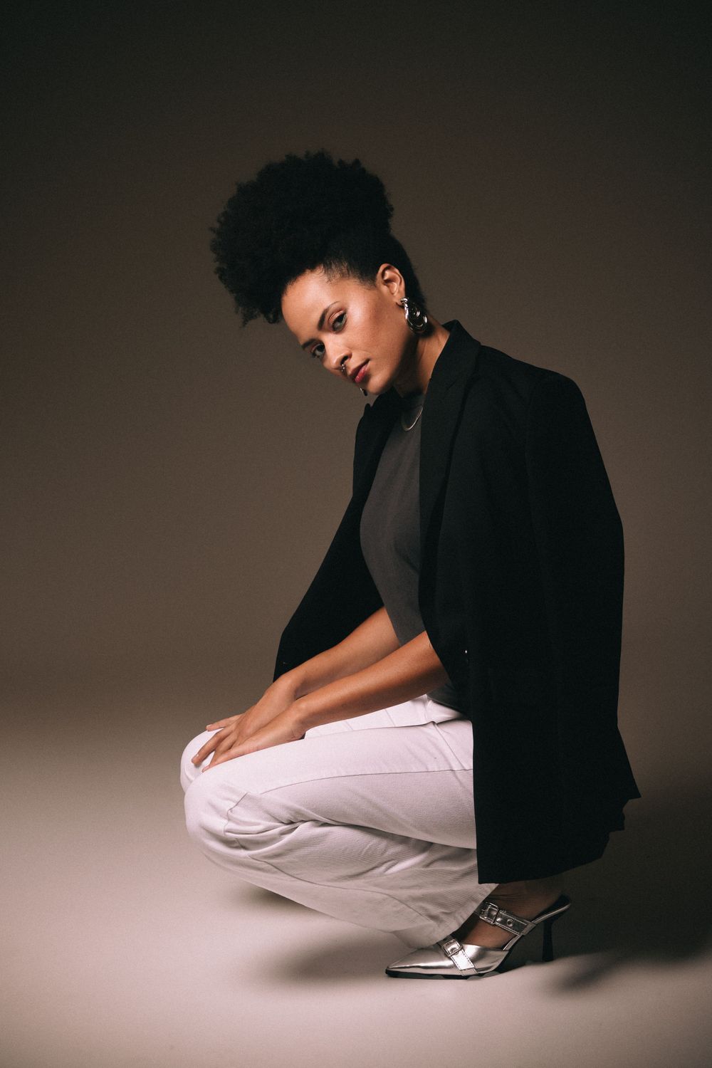Fashion model in black blazer and white pants crouches in dramatic pose against brown backdrop.