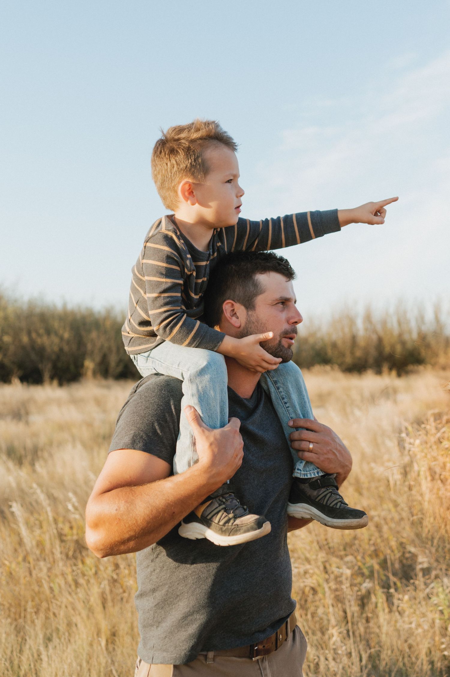 Child sitting on parent's shoulders pointing at something in a sunlit field.