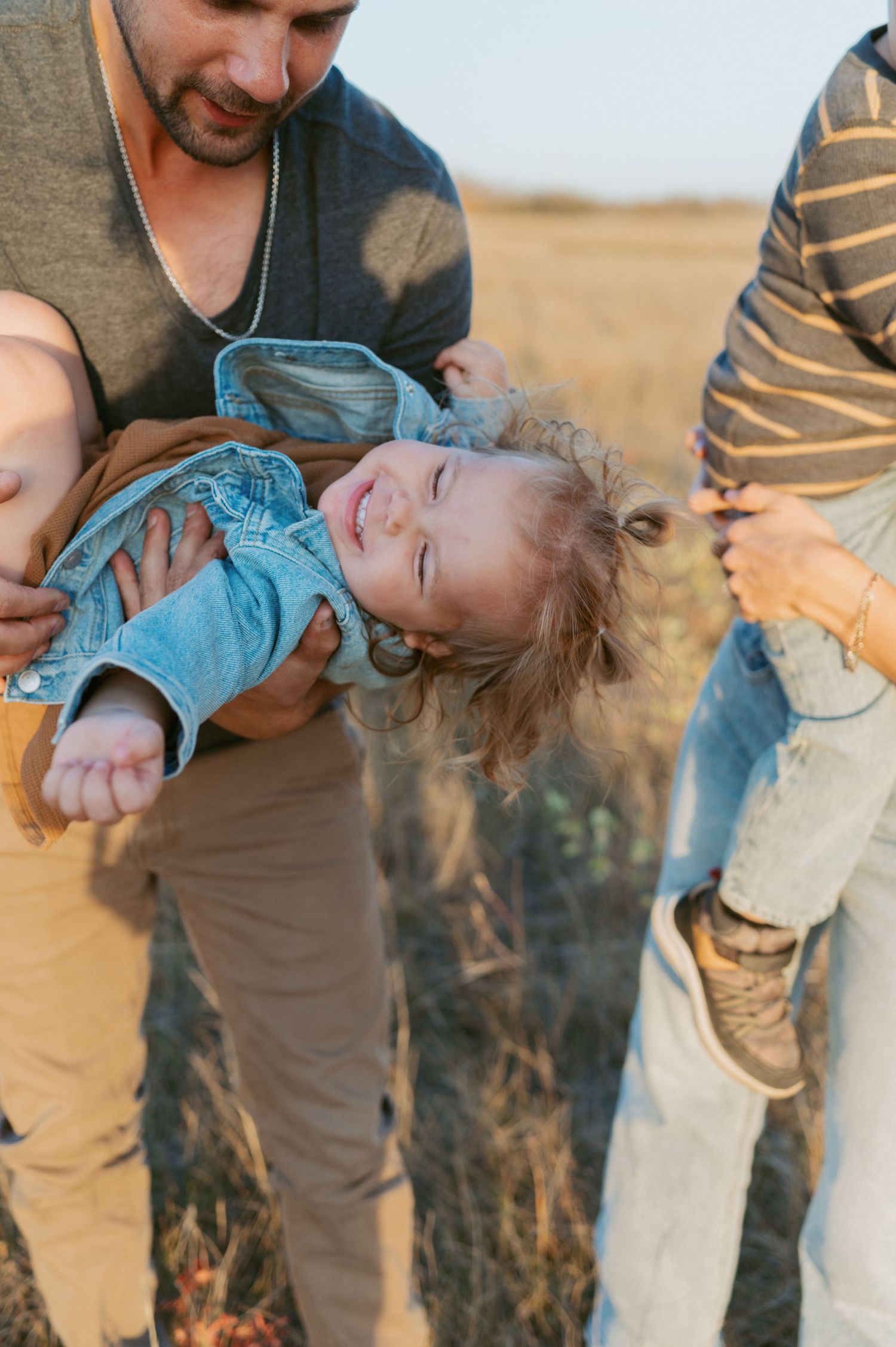 A playful family moment of parental figures holding and swinging a small child in denim against a desert landscape.