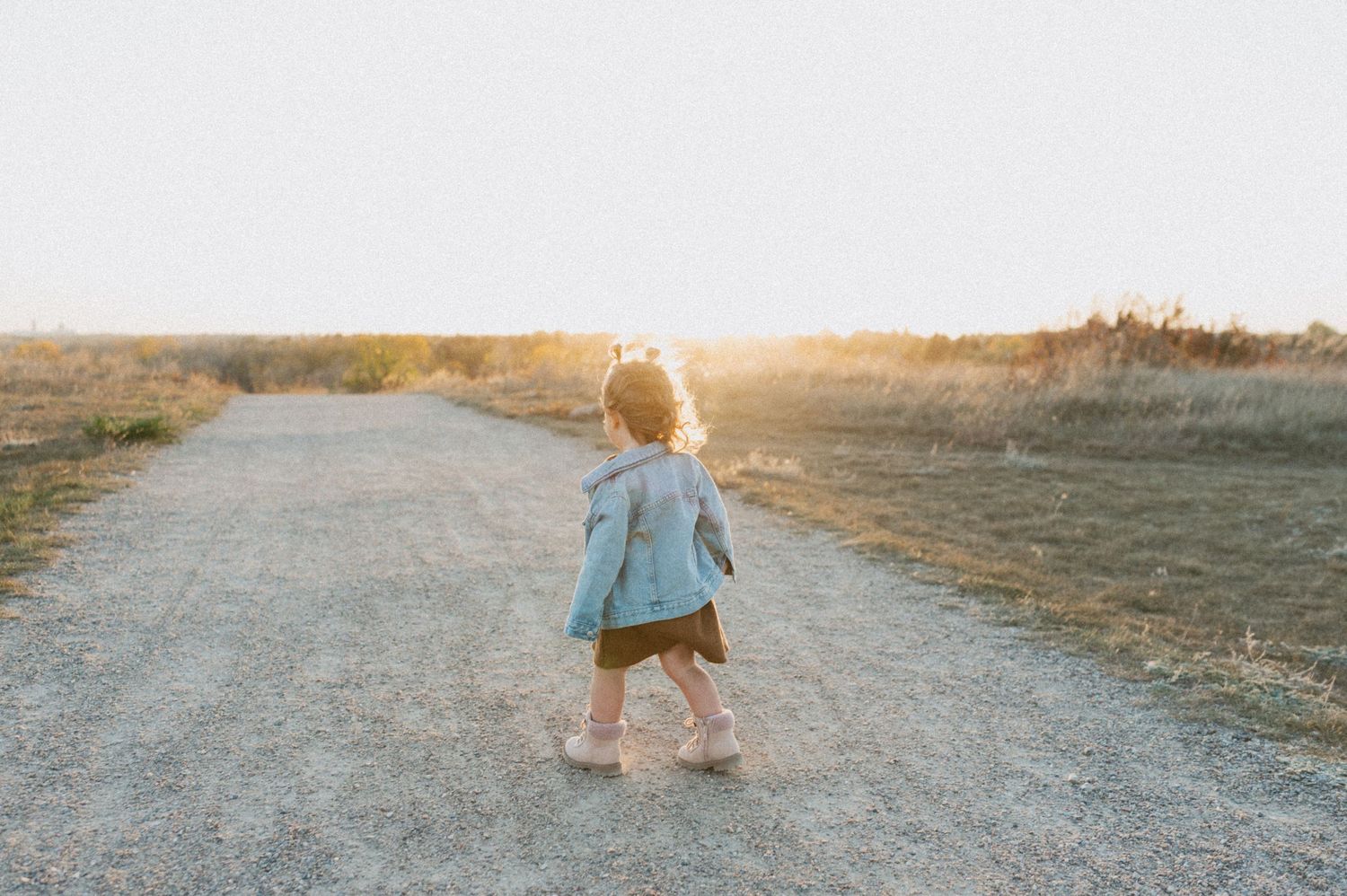 Small toddler in denim walking alone on a dirt path during golden hour.