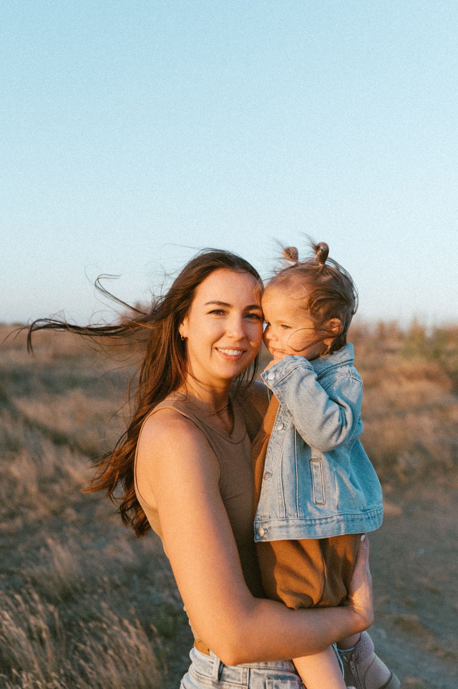 Joyful outdoor portrait of parent and toddler with wind-blown hair at sunset.