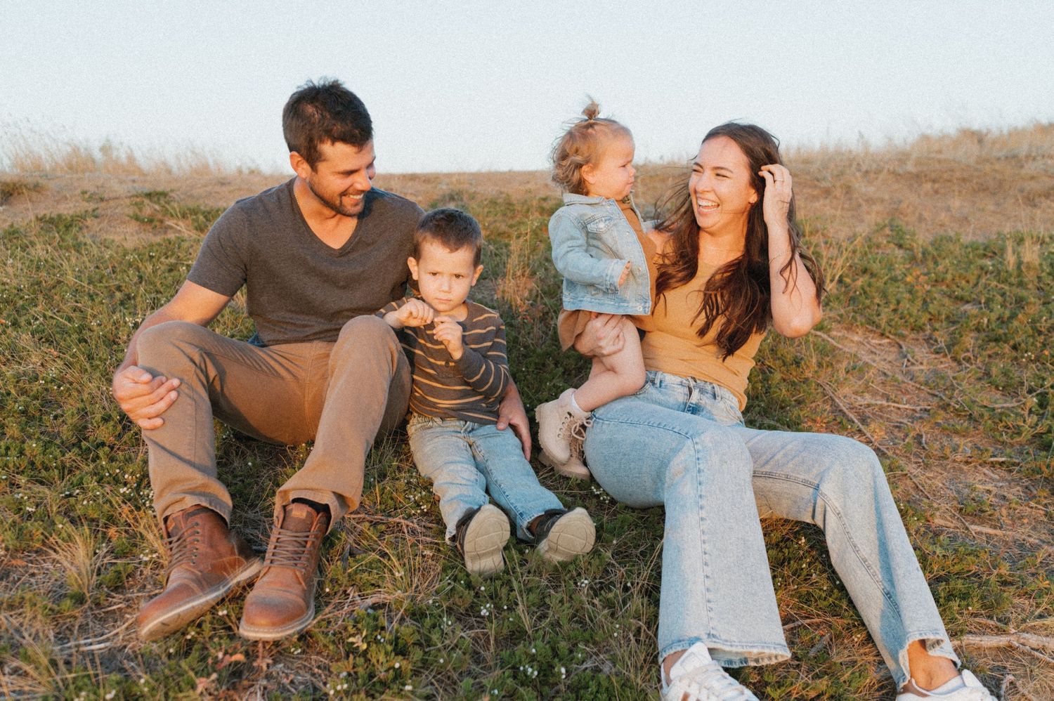 Family of four sitting together on grassy hill during sunset photoshoot.