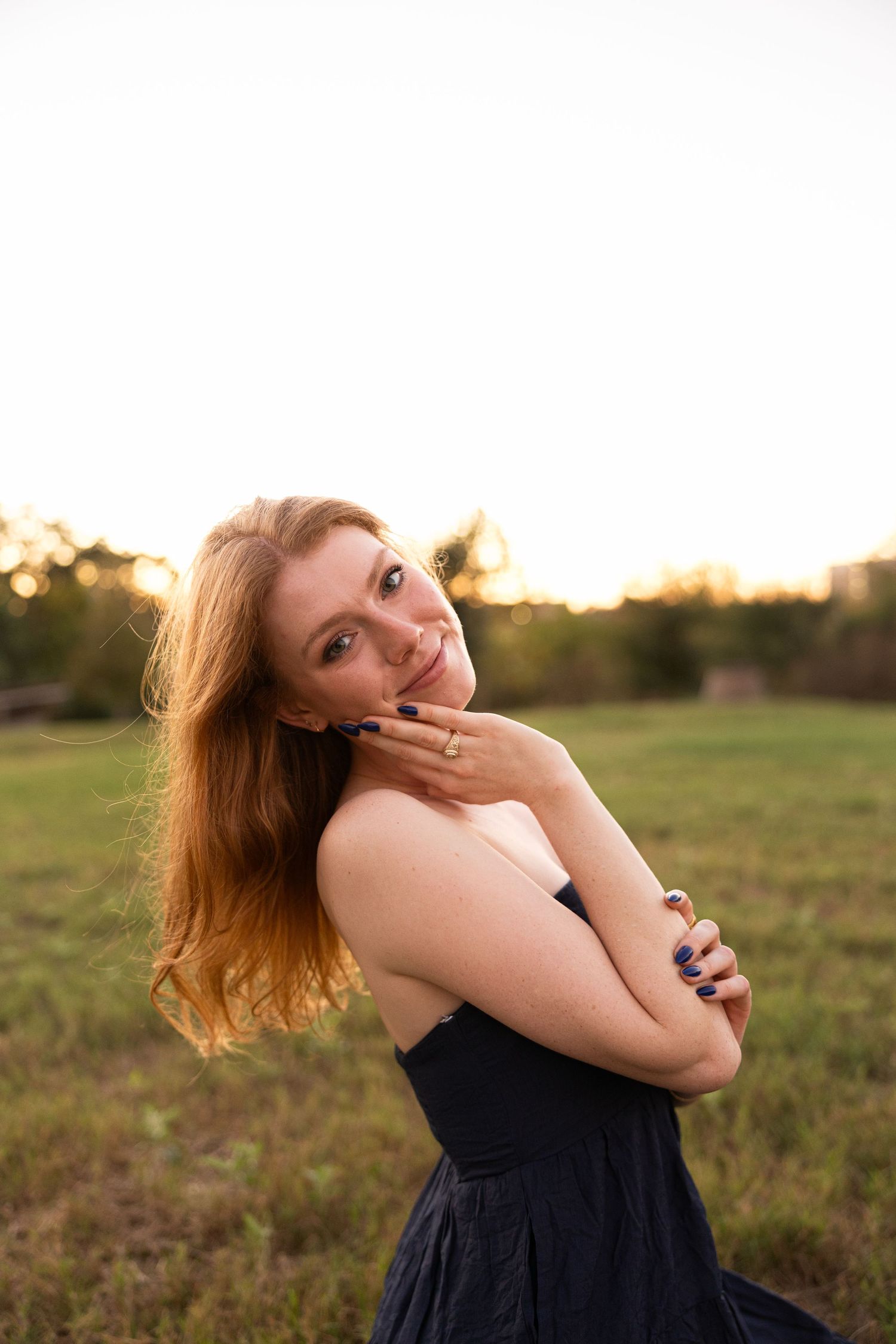 A person in a black dress poses playfully in a sunlit field during golden hour.