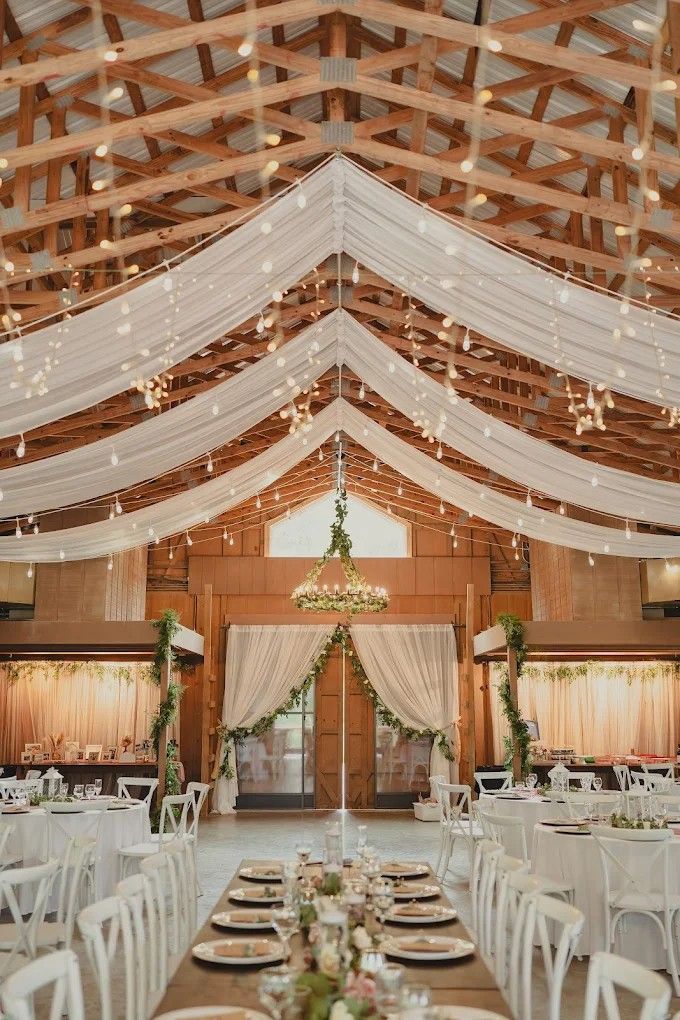 Barn-style wedding venue with white drapes, string lights, and exposed wooden beams creating a romantic atmosphere.