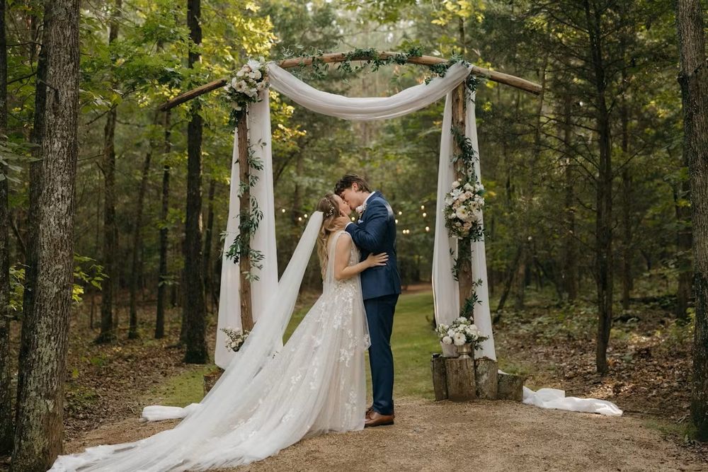 A couple shares a romantic moment under a rustic white fabric arch decorated with flowers in a wooded forest setting.