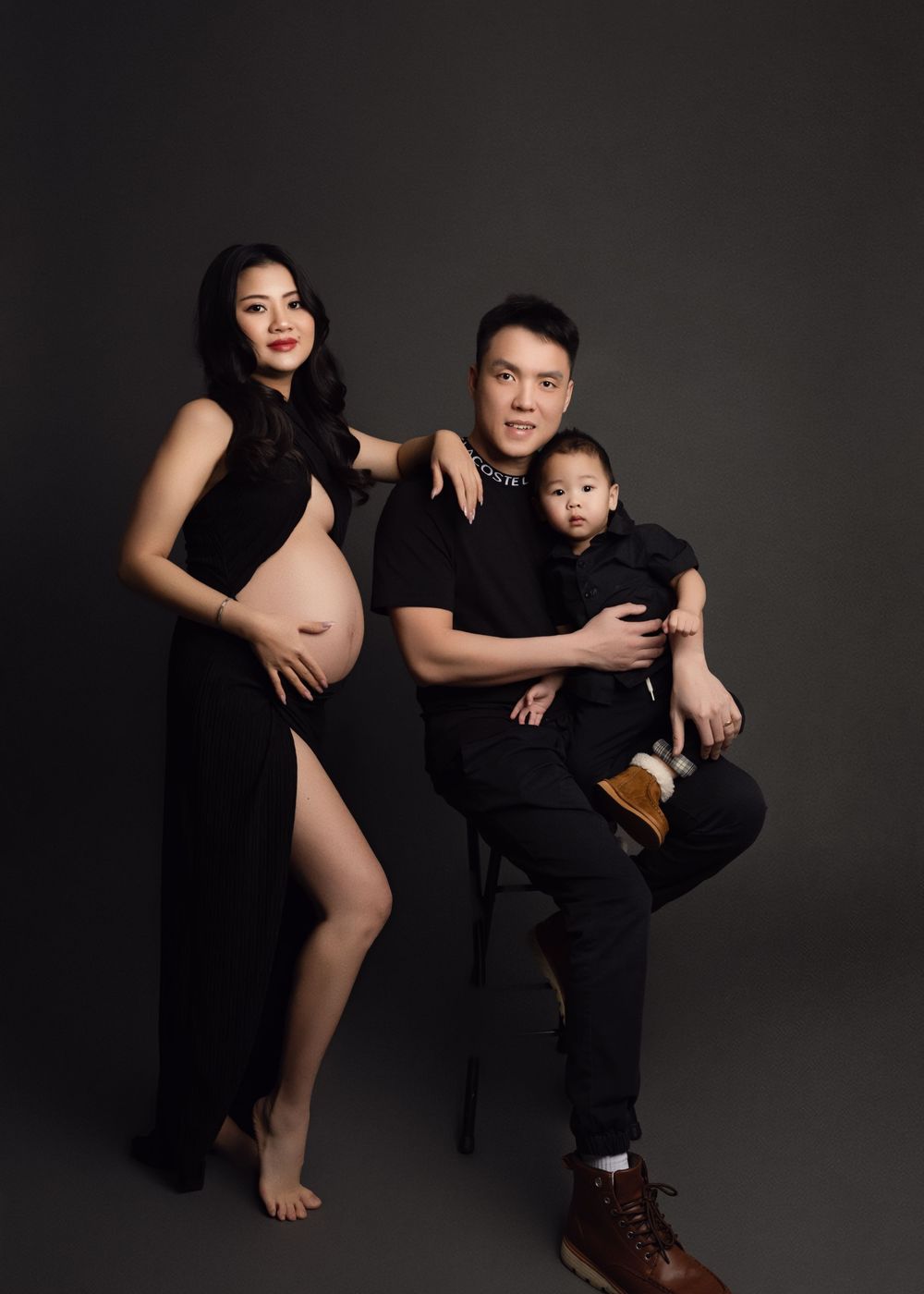 Artistic maternity portrait featuring family of three in black outfits against dark backdrop in studio setting.