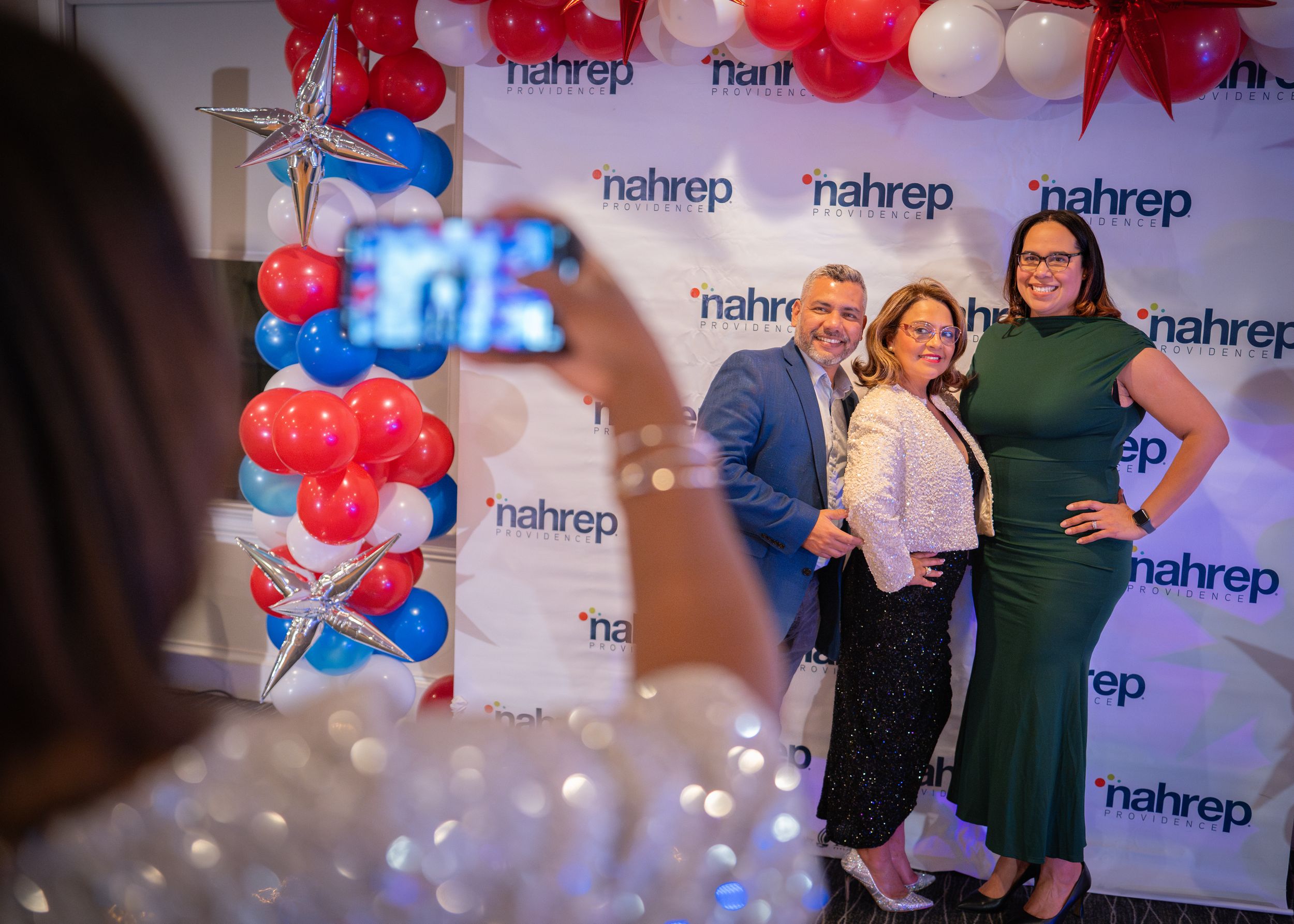 People gather at a red and white balloon decorated event with a blue and white branded backdrop.