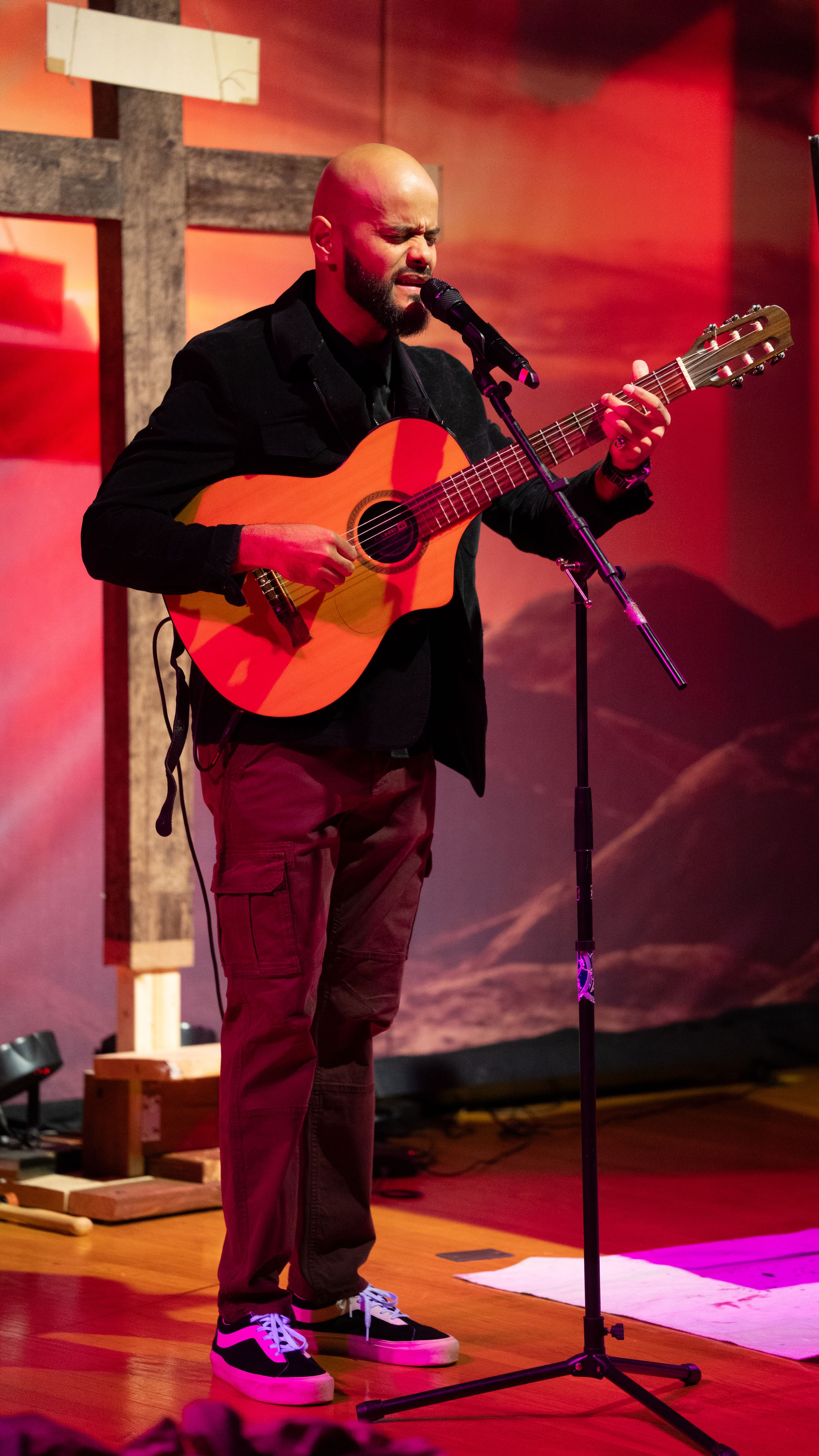 Musician performs on acoustic guitar while standing at microphone on stage with dramatic red lighting.