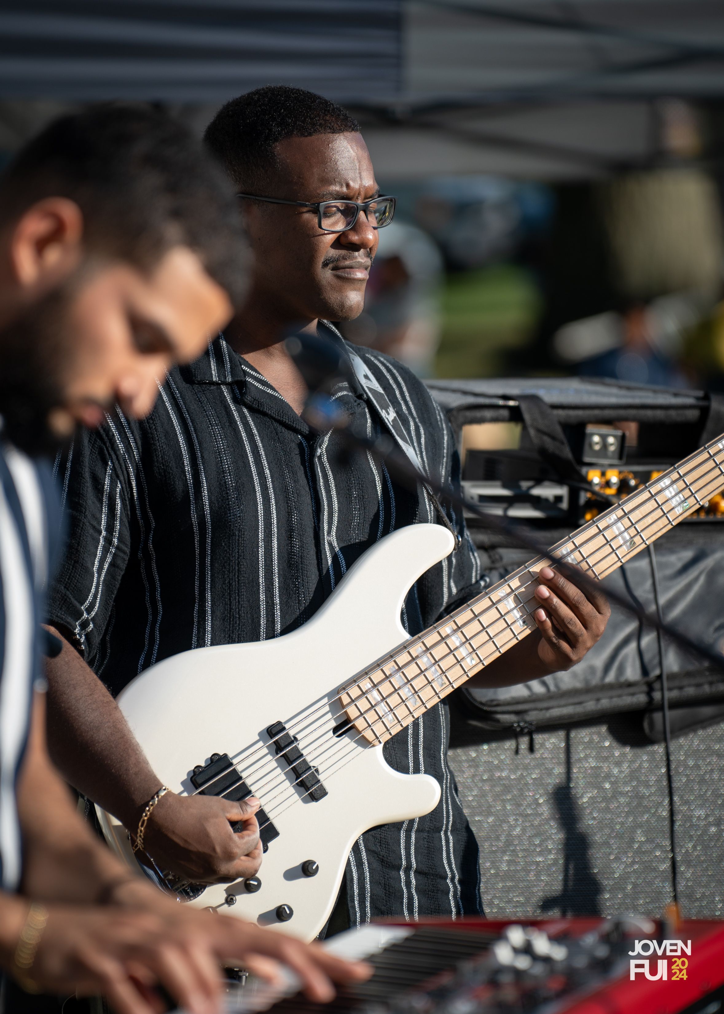 Musicians perform together at an outdoor event with one playing a white bass guitar while wearing glasses and a striped shirt.