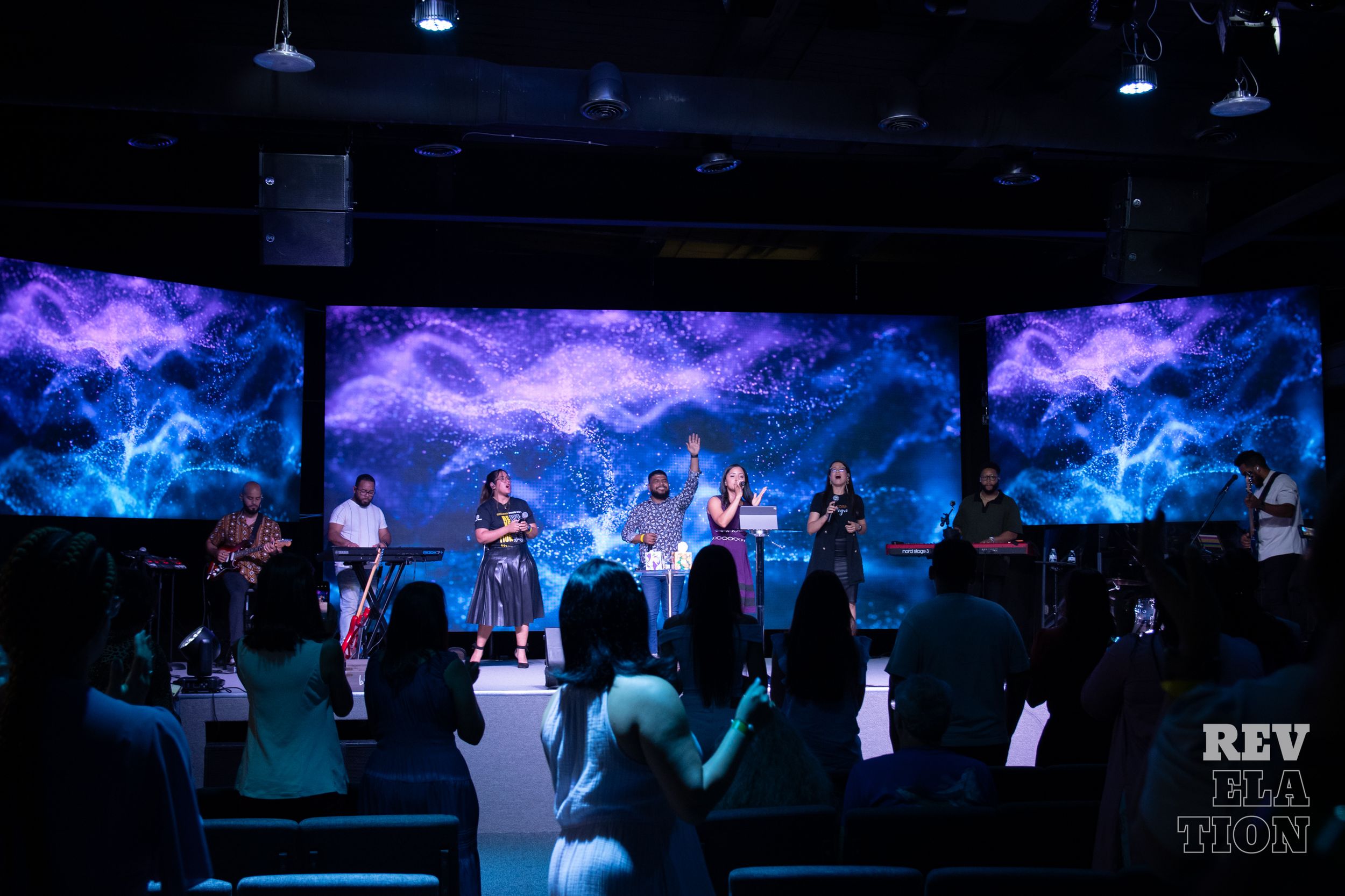 Musicians perform on stage against a backdrop of vibrant blue and purple digital lightning effects at a concert.
