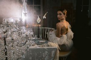 An elegant dinner setting with crystal chandeliers and candelabras casts a romantic glow as someone poses at a formal table.