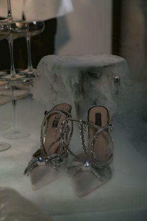 Elegant crystal-embellished silver bridal shoes with wine glasses and dry ice creating a misty ambiance on a white surface.