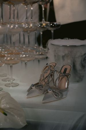 A set of elegant silver evening shoes with crystal embellishments displayed alongside wine glasses on a white surface.