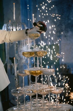 Champagne being poured into a tower of coupe glasses with sparkling lights in the background.