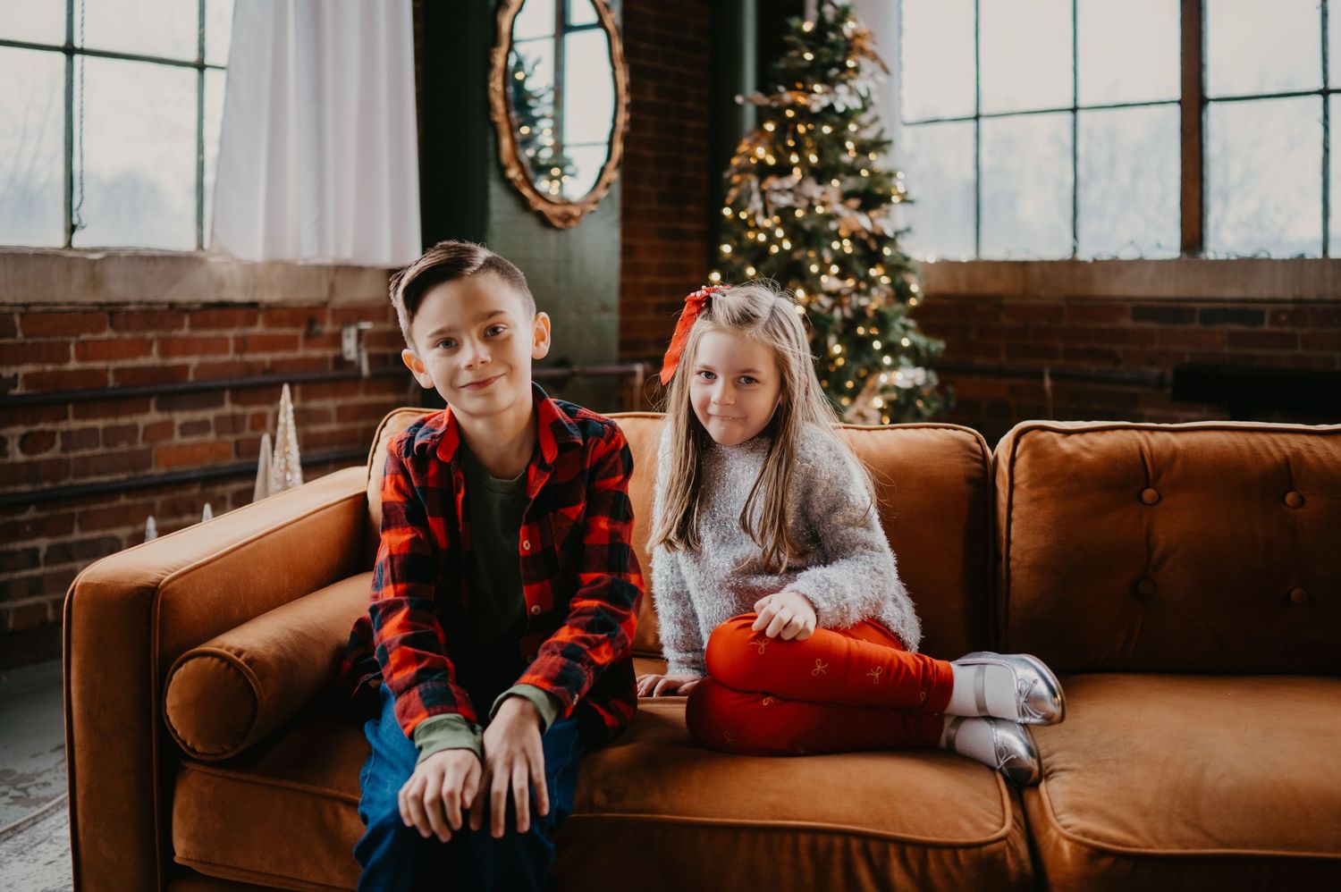 Two children sitting on a brown leather couch in front of a decorated Christmas tree in a festive holiday setting.