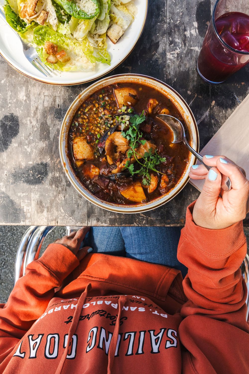 Overhead view of hearty stew with vegetables and red wine served alongside a Caesar salad on dark table.