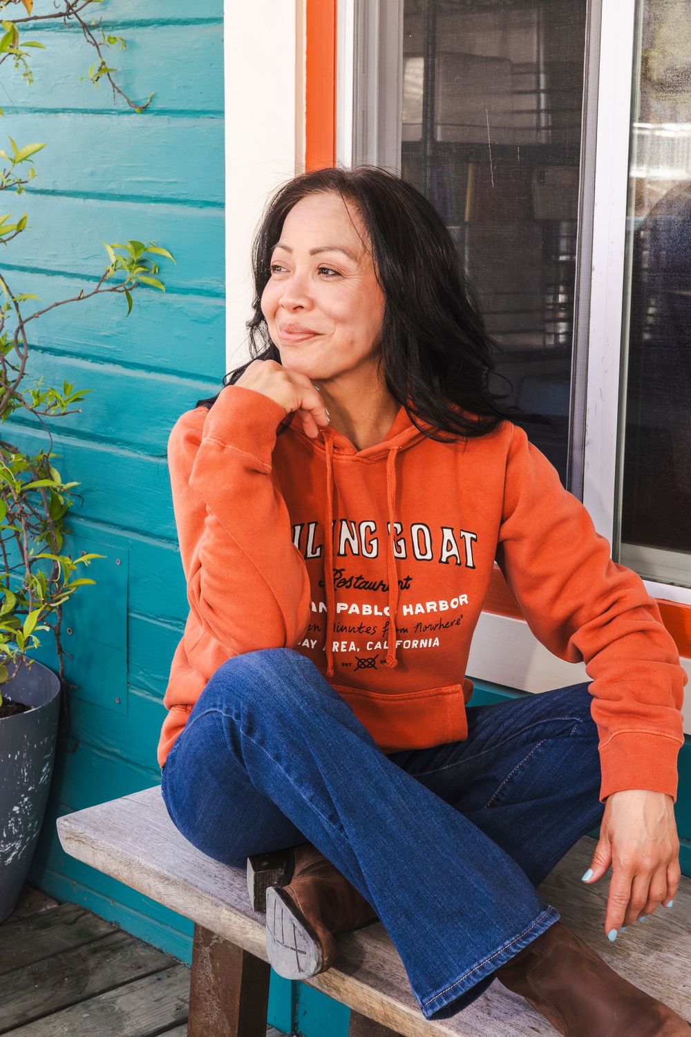 Someone wearing an orange Flying Goat hoodie and blue jeans sits casually on a bench against a turquoise wall.