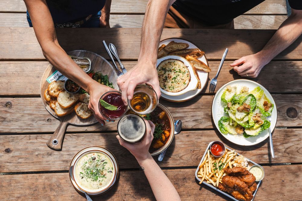 Overhead view of people sharing drinks and food including salad, fries and dips on a rustic wooden table.