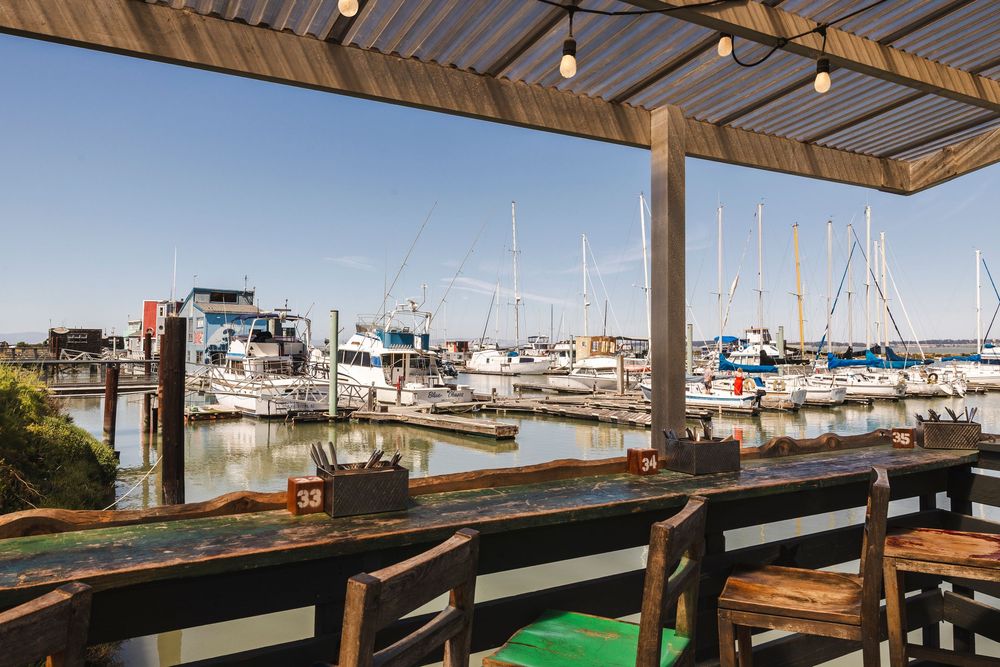 Wooden deck overlooking a marina with boats and houseboats docked under blue skies at a waterfront restaurant.