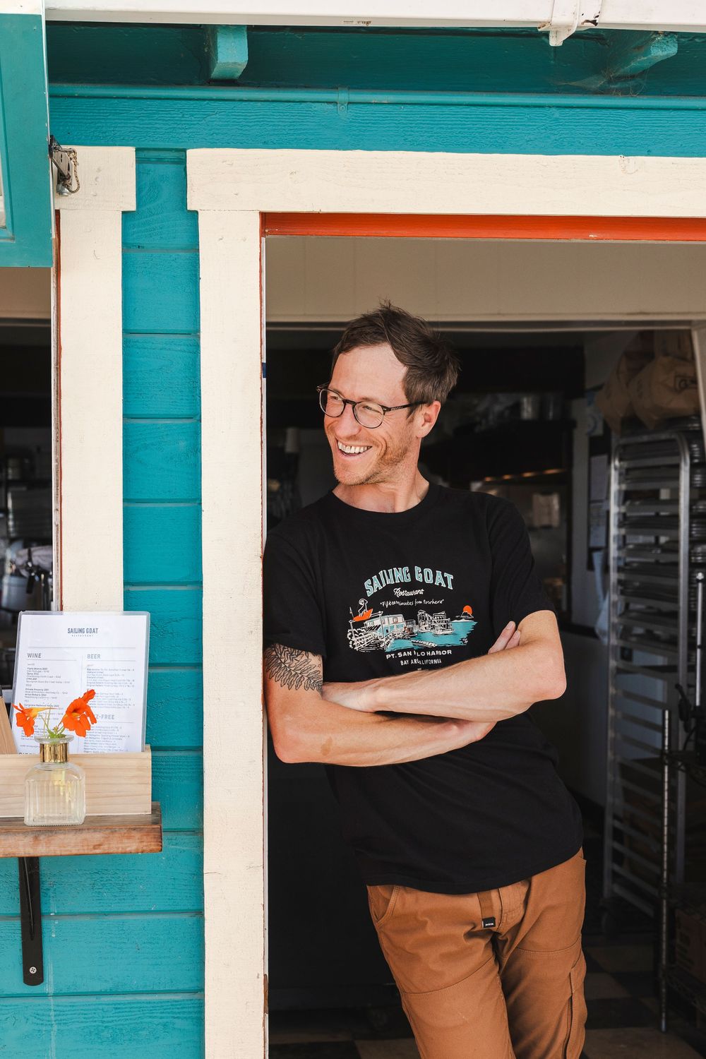Person wearing glasses and black t-shirt smiling through service window of turquoise-colored building.