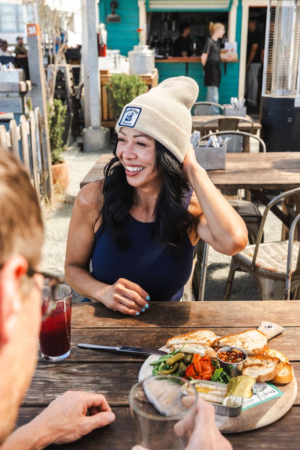 People enjoying casual outdoor dining at a restaurant patio with plates of food and drinks on wooden tables.