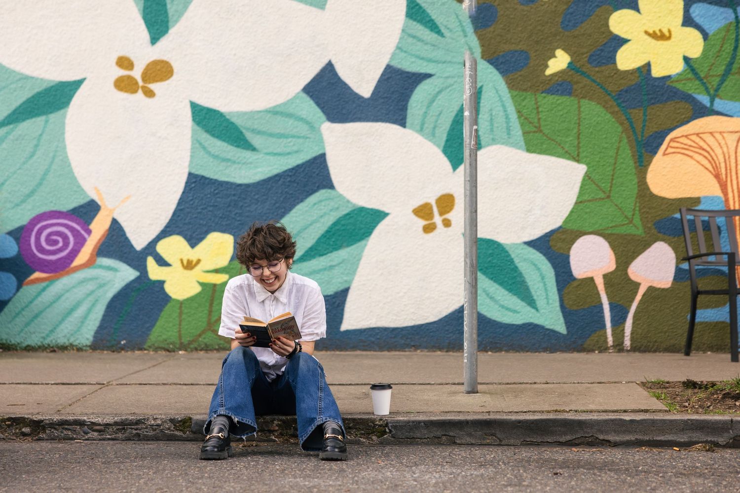 A colorful floral mural with white and orange flowers adorns an outdoor wall while someone sits on a ledge below.