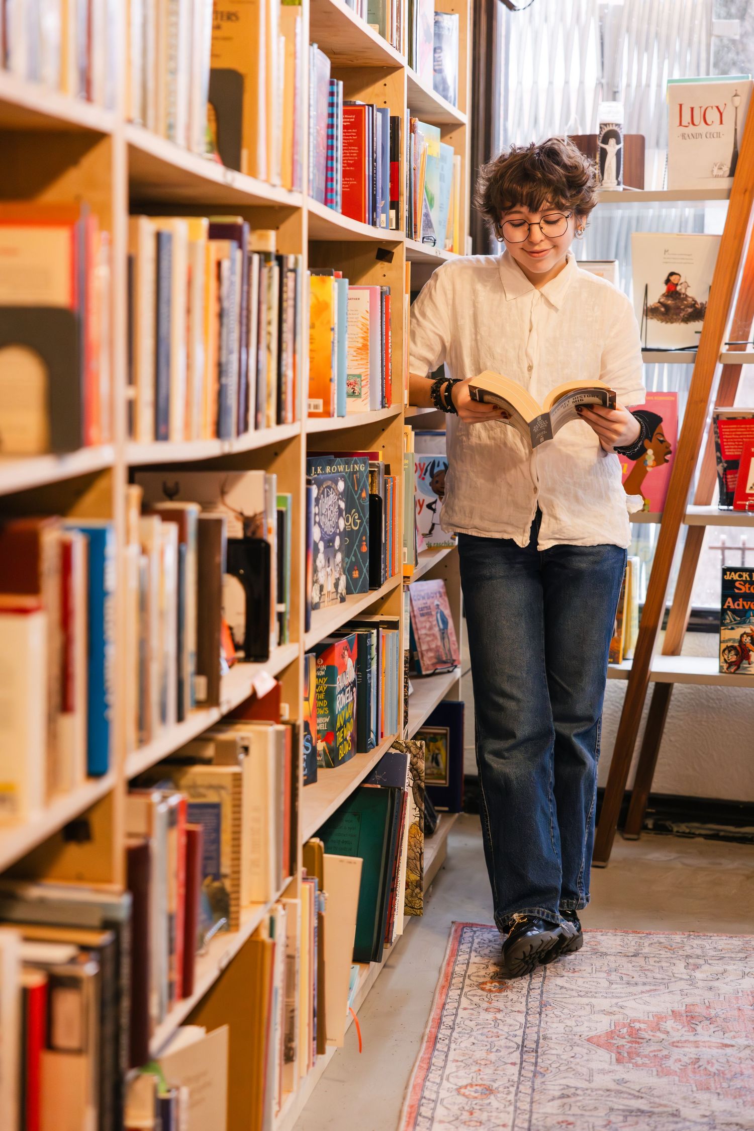 Someone browses books while standing in an aisle between tall wooden bookshelves in a library or bookstore.