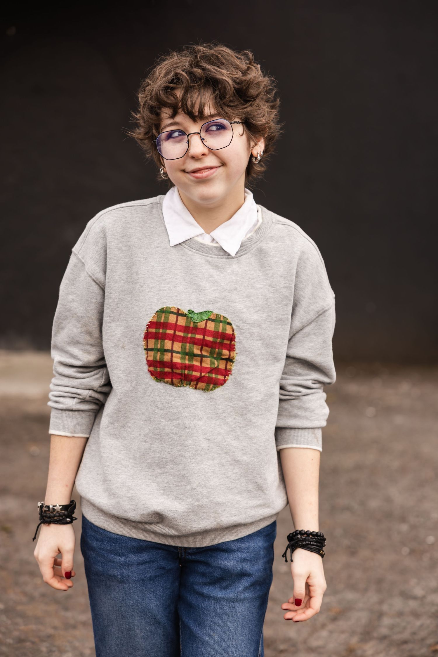 A person wearing a cozy gray sweater with plaid tartan heart detail and blue jeans against a dark background.
