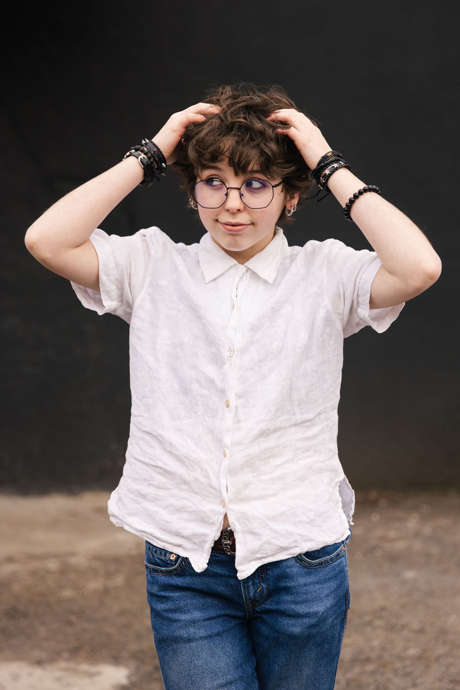 A person in round glasses wearing a white button-down shirt and jeans runs hands through curly hair against a dark background.